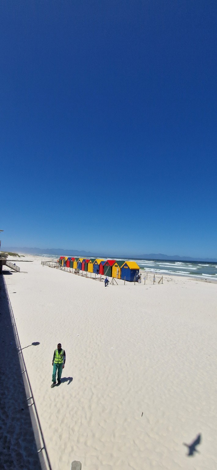 Wide view of Muizenberg Beach with white sand, the distant row of colourful Victorian bathing huts and the False Bay shoreline, Cape Town