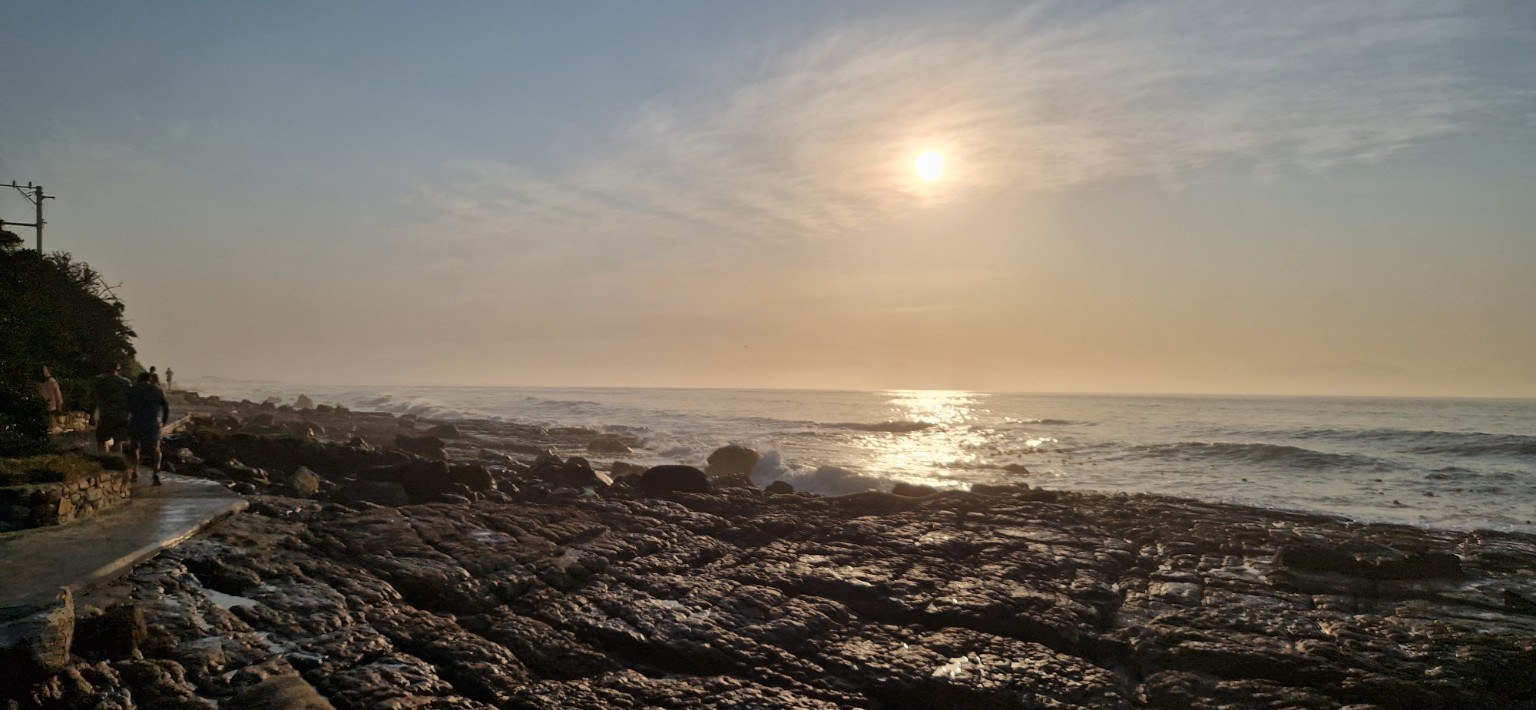 Muizenberg sunrise over False Bay Golden sunrise light over the rocky False Bay coastline at Muizenberg with walkers along the shoreline path, Cape Town