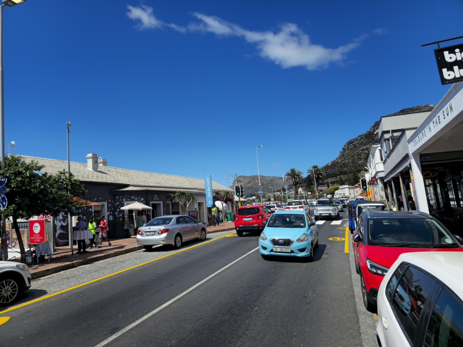 Kalk Bay Main Road on a sunny day with the Kalk Bay Station sign, boutique shops, restaurants and mountain backdrop, Cape Town
