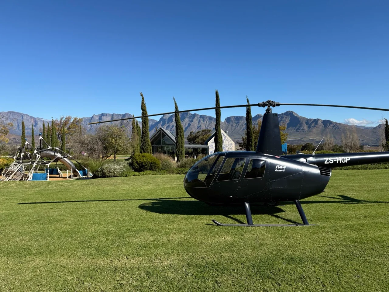 Black Robinson R44 Raven II helicopter, registration ZS-HGP, standing on a manicured lawn with cypress trees and mountain peaks behind it under a clear blue sky