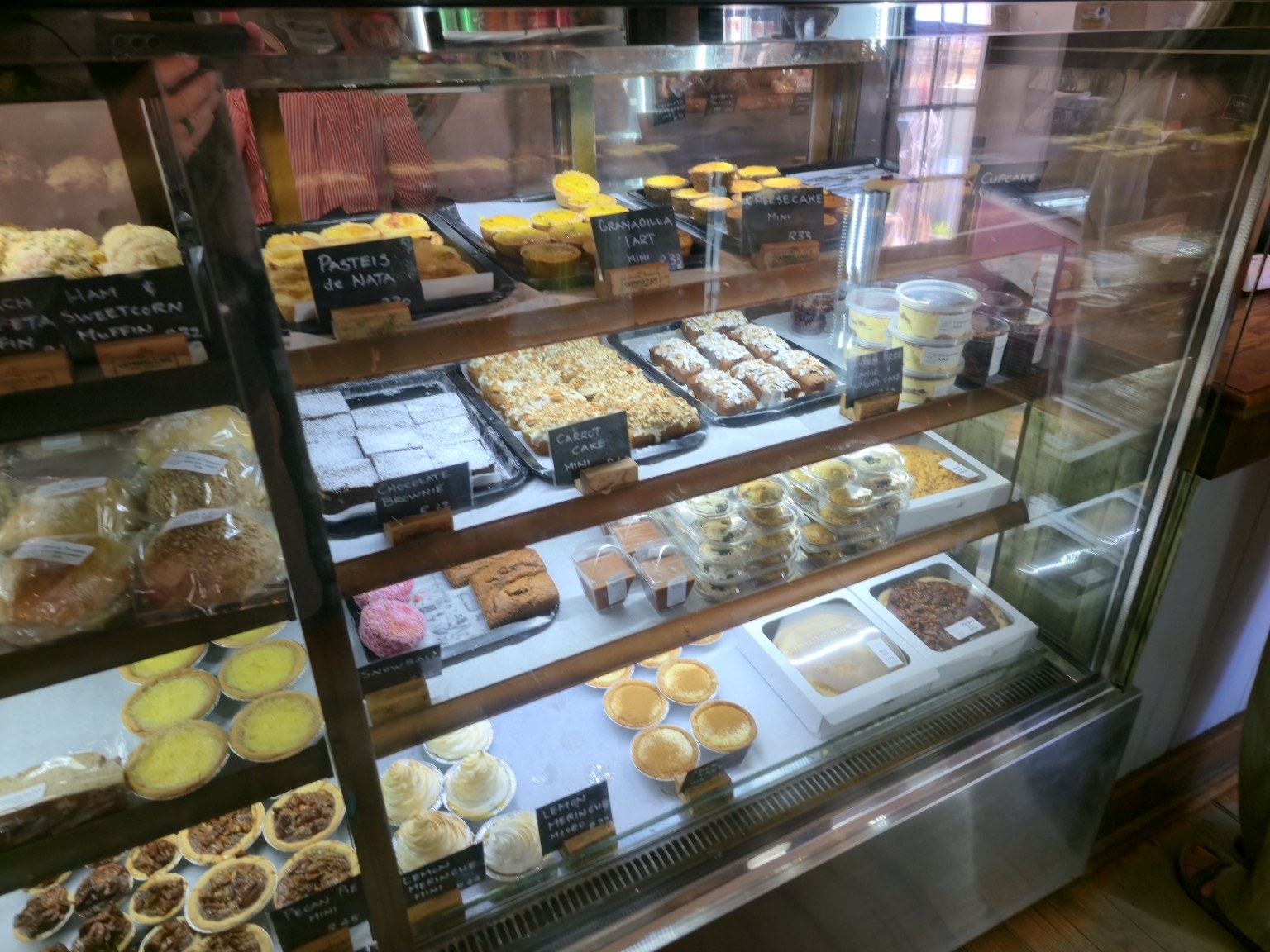 Glass display cabinet at Olympia Café & Deli on Main Road Kalk Bay filled with pasteis de nata, carrot cake, lemon meringue tarts, pecan tarts and freshly baked pastries, Cape Town
