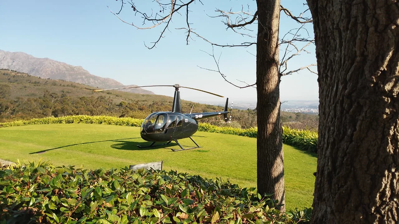 Black Robinson R44 helicopter parked on a grass landing area with mountains and fynbos-covered hills in the background