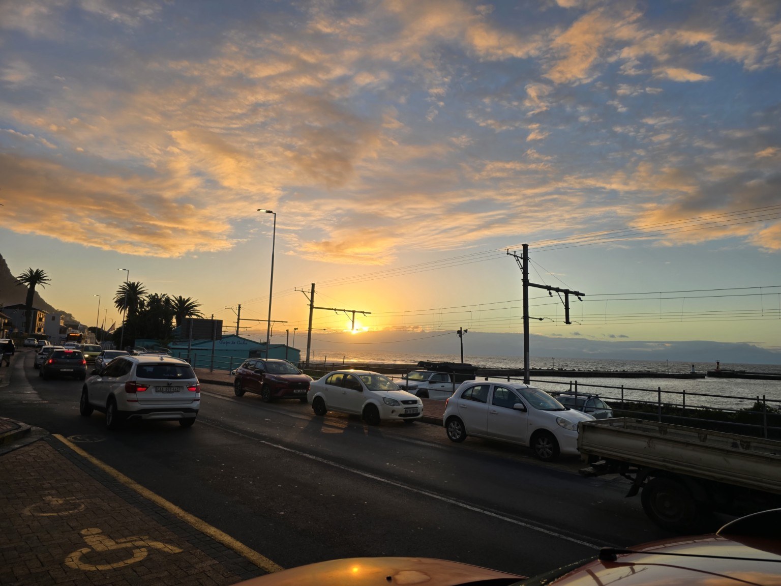 Golden sunset over False Bay from Kalk Bay Main Road, with fishing boats along the harbour wall and Muizenberg Mountain silhouetted against the sky