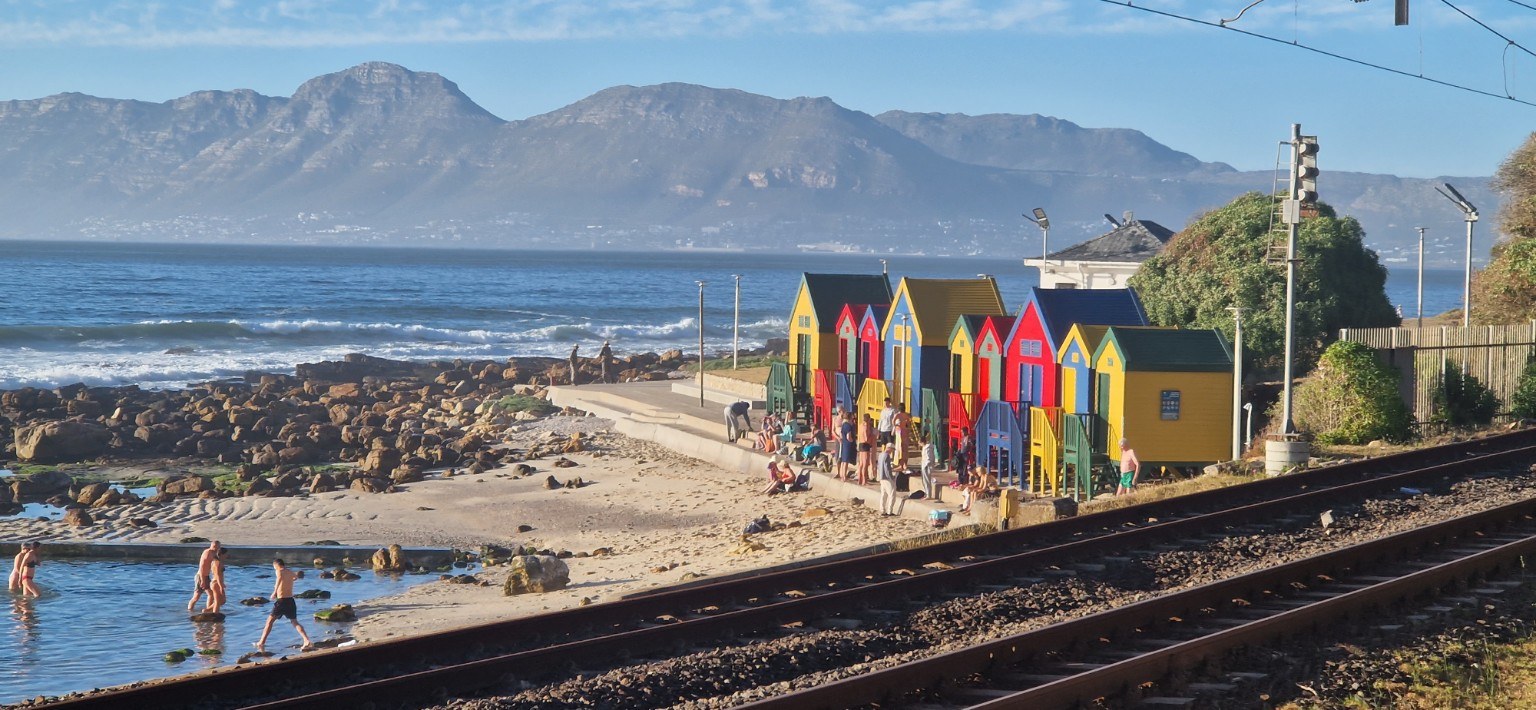 The famous colourful St James bathing boxes beside the sheltered tidal pool, Southern Line railway and Muizenberg Mountain, False Bay, Cape Town